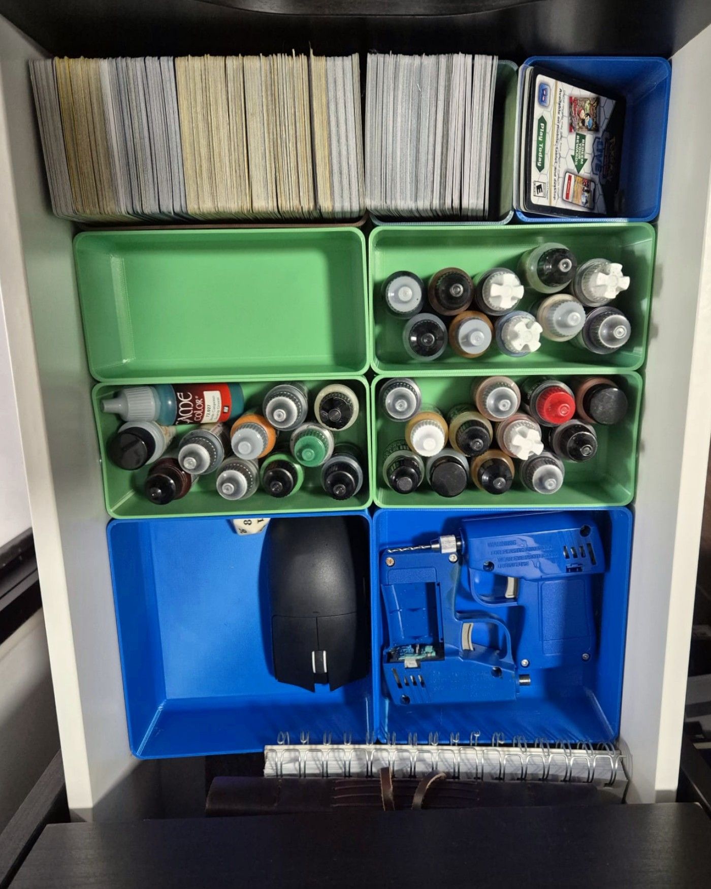 Drawer organised with green and blue 3D-printed bins holding model paints, trading cards, and tools